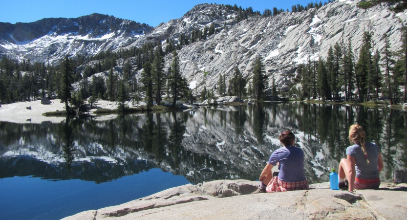 two students sit on a rock facing a blue alpine lake reflecting a mountainous landscape